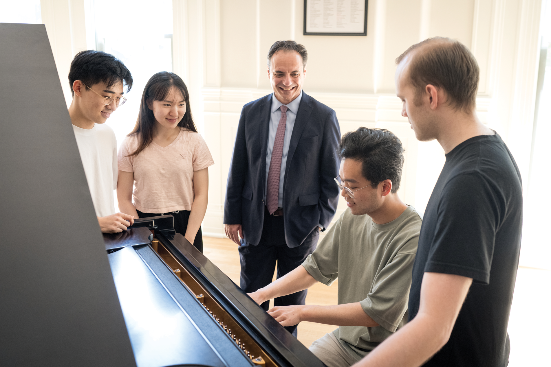 Four students standing around a piano, one student sitting, and man standing, smiling